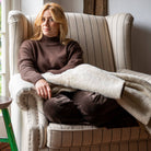 A woman sits in a cream wingback armchair, wrapped in a light beige throw blanket, wearing a brown turtleneck sweater and dark brown pants in a softly lit indoor setting.