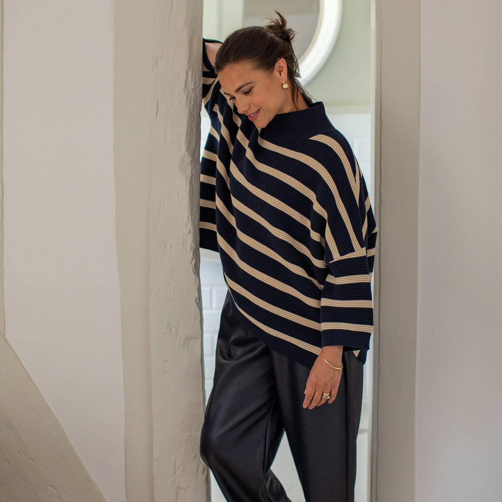 A woman wearing a navy and beige striped knit sweater and dark wide-leg pants, standing barefoot in a doorway with white walls.