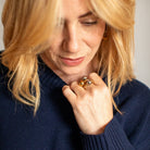 A close-up lifestyle shot of a woman with blonde hair wearing a dark navy blue ribbed knit sweater and a gold ring with dark stones.