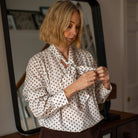 A woman with blonde hair wearing a white long-sleeve blouse with a brown polka dot pattern and a ruffled tie-neck detail, paired with brown corduroy pants, standing in front of a large mirror.