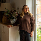 A woman wearing a brown ribbed turtleneck sweater and dark leather pants stands indoors next to a window, with a cabinet, lamp, and dried flowers in the background.