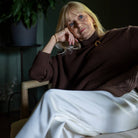 A woman with blonde hair sits in a dark armchair, wearing a dark brown ribbed turtleneck sweater with a gold button detail and white trousers.