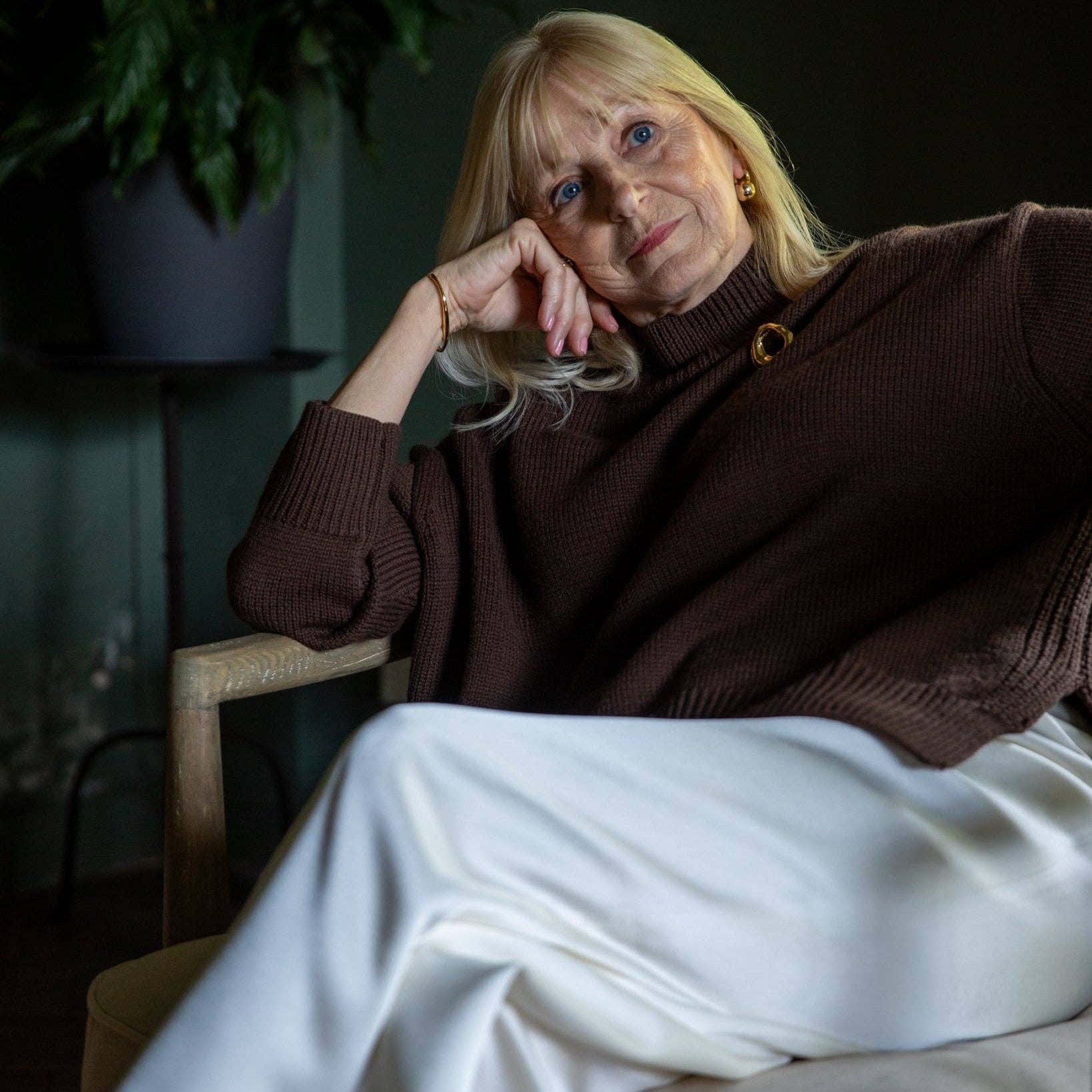 A woman with blonde hair sits in a dark armchair, wearing a dark brown ribbed turtleneck sweater with a gold button detail and white trousers.