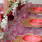Decorative table setting with pink plates, glasses, and flowers on a patterned tablecloth.