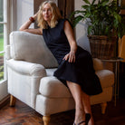 Woman in a black dress sitting on a beige armchair in a room with plants and a window.