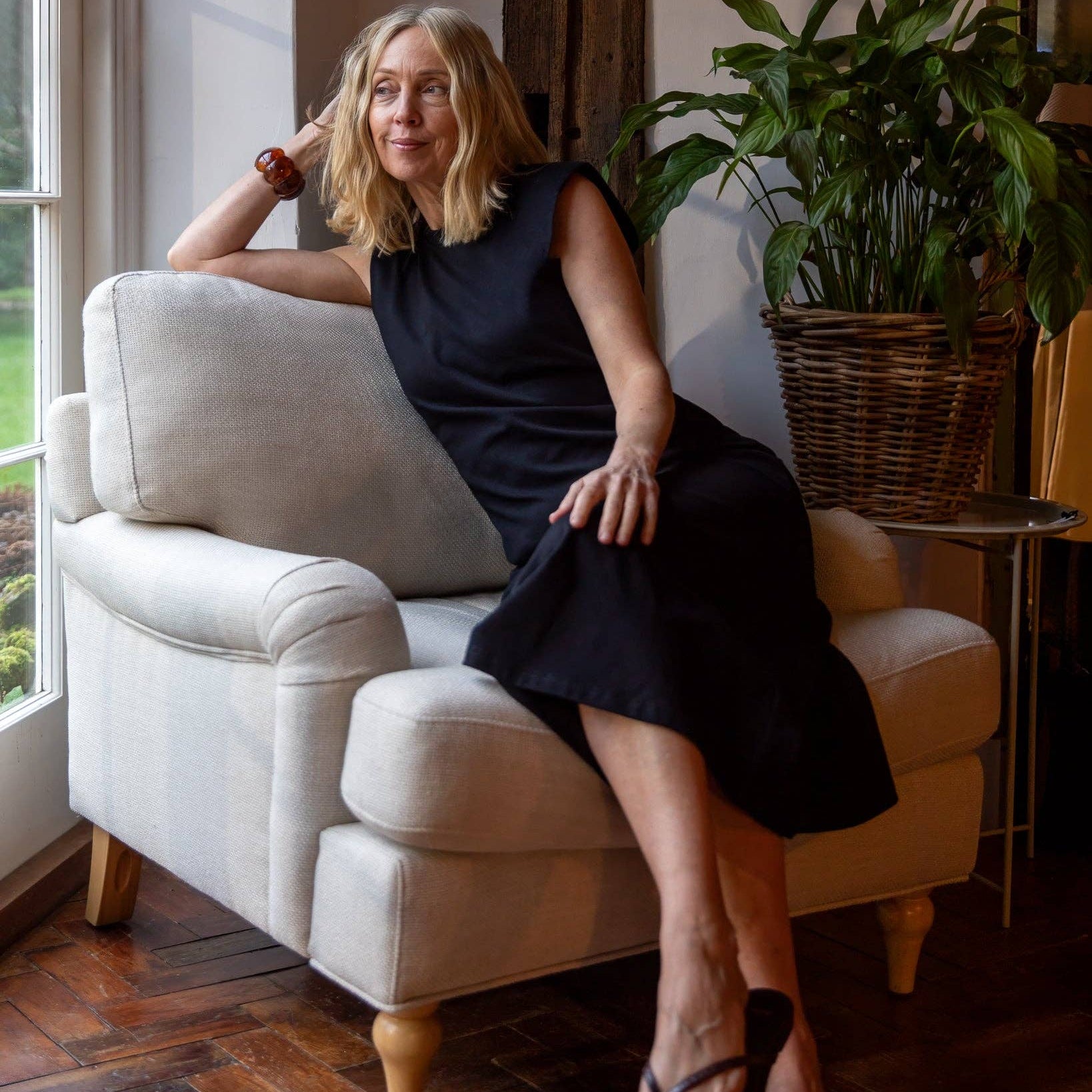 Woman in a black dress sitting on a beige armchair in a room with plants and a window.