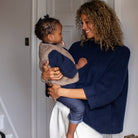A woman with curly brown hair holds a baby in a home setting. The woman is wearing a dark navy blue knit sweater and white trousers, while the baby wears a beige and navy blue knit outfit.