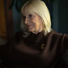 A close-up portrait of a woman with blonde hair wearing a dark brown ribbed turtleneck sweater with a gold crescent moon pin on the chest, set against a dark, moody background.