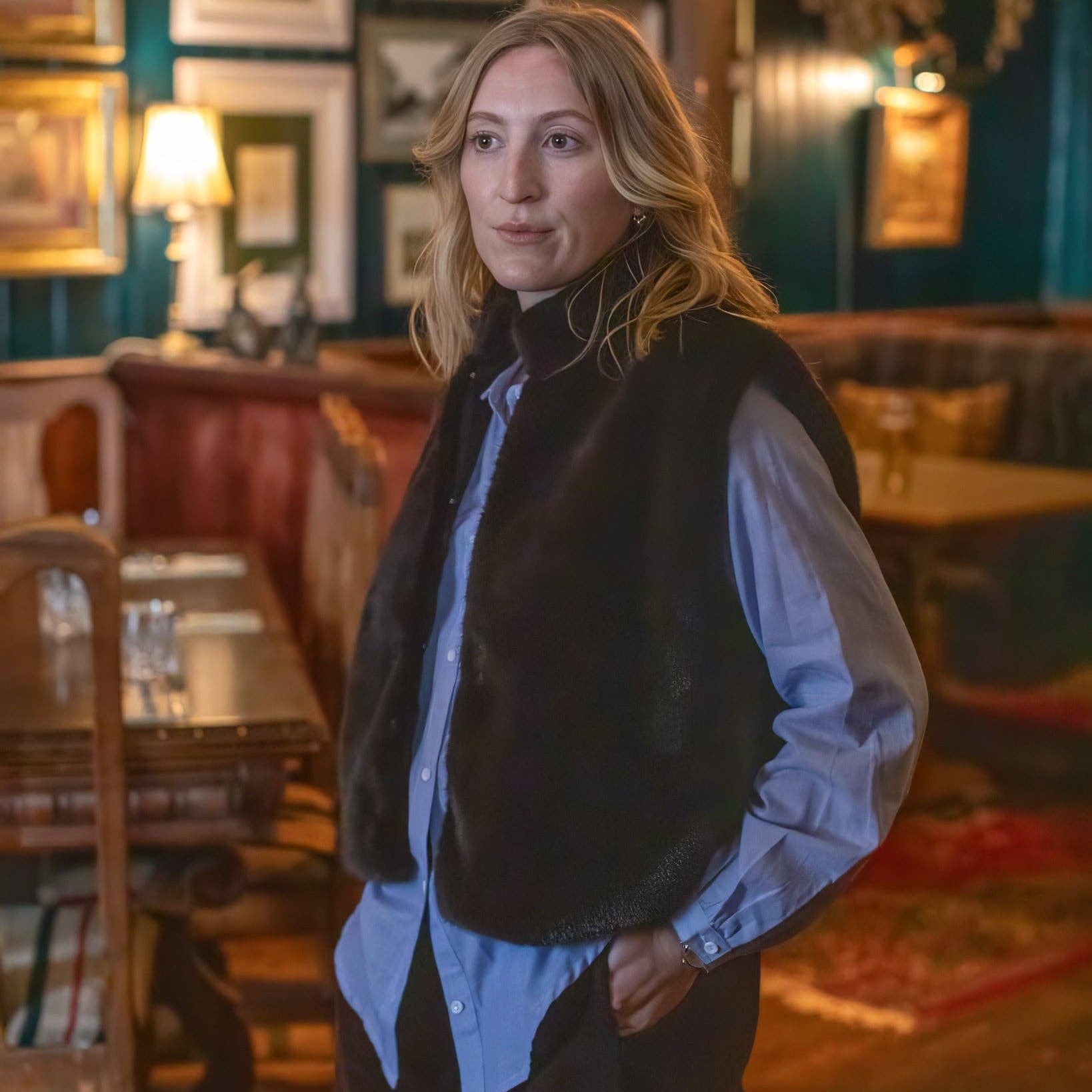A woman standing in a dimly lit, vintage-style pub or cafe, wearing a brown faux-fur vest over a light blue button-down shirt and dark trousers.