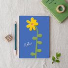 Notebook with a yellow flower illustration on a blue background, placed on a textured surface with stationery items.