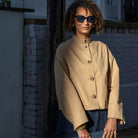 Person wearing a beige coat and sunglasses, holding a blue bag, standing against a wall.