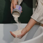 Person pouring a white powder from a purple container into their hand over a white surface.