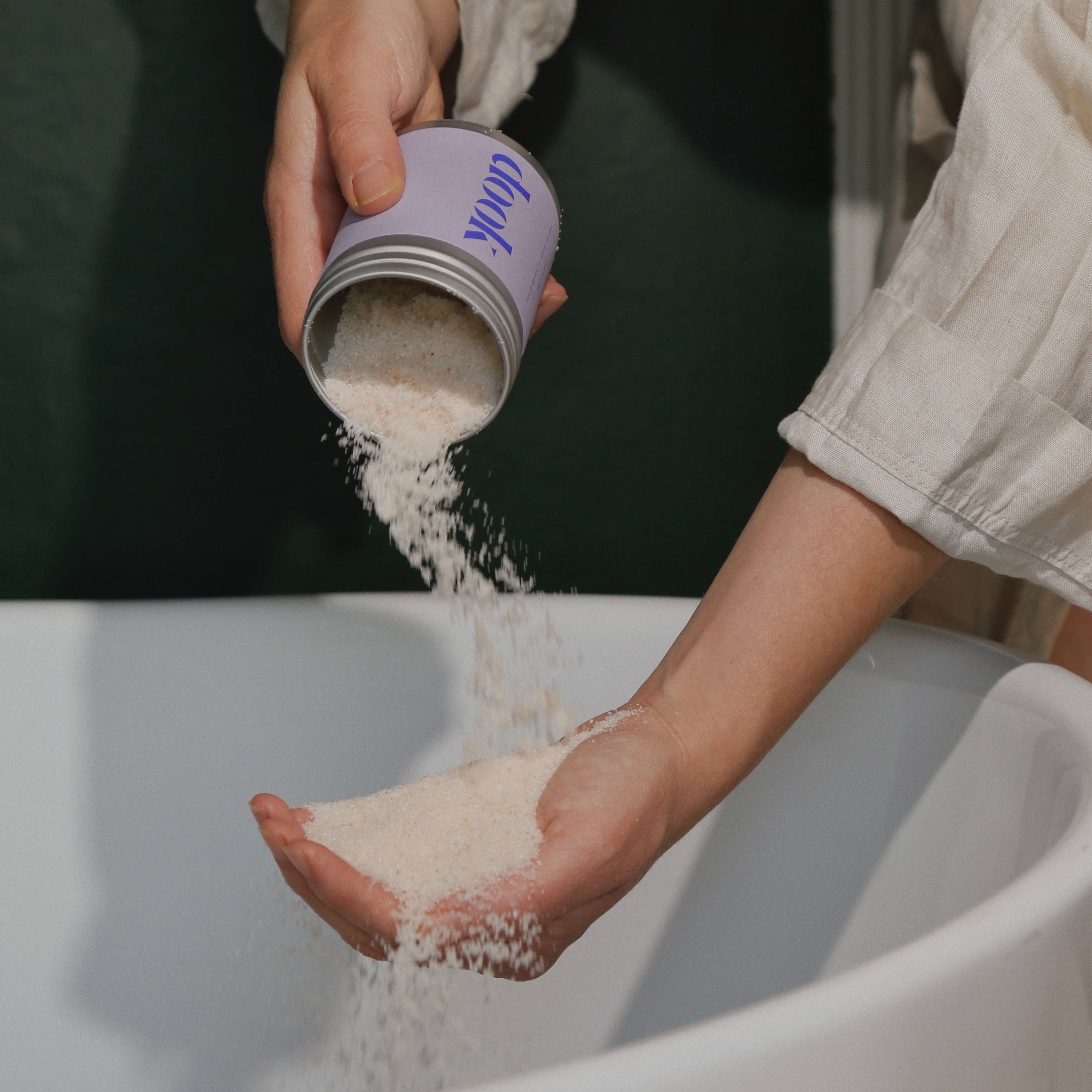 Person pouring a white powder from a purple container into their hand over a white surface.