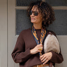 Woman wearing a brown sweater with fur lining, sunglasses, and holding a bag against a neutral background.