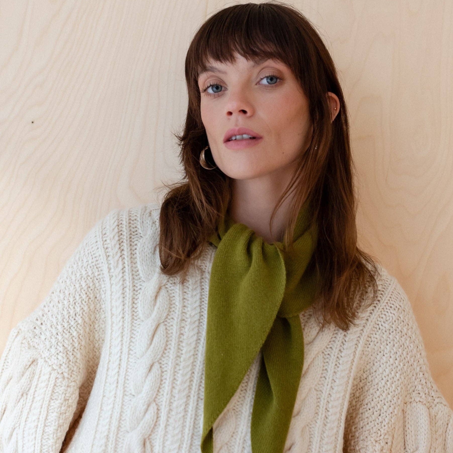 Woman wearing a white cable knit sweater with a green scarf against a beige background