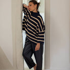 A woman stands barefoot in a doorway wearing a navy and beige striped knit sweater and dark wide-leg pants.