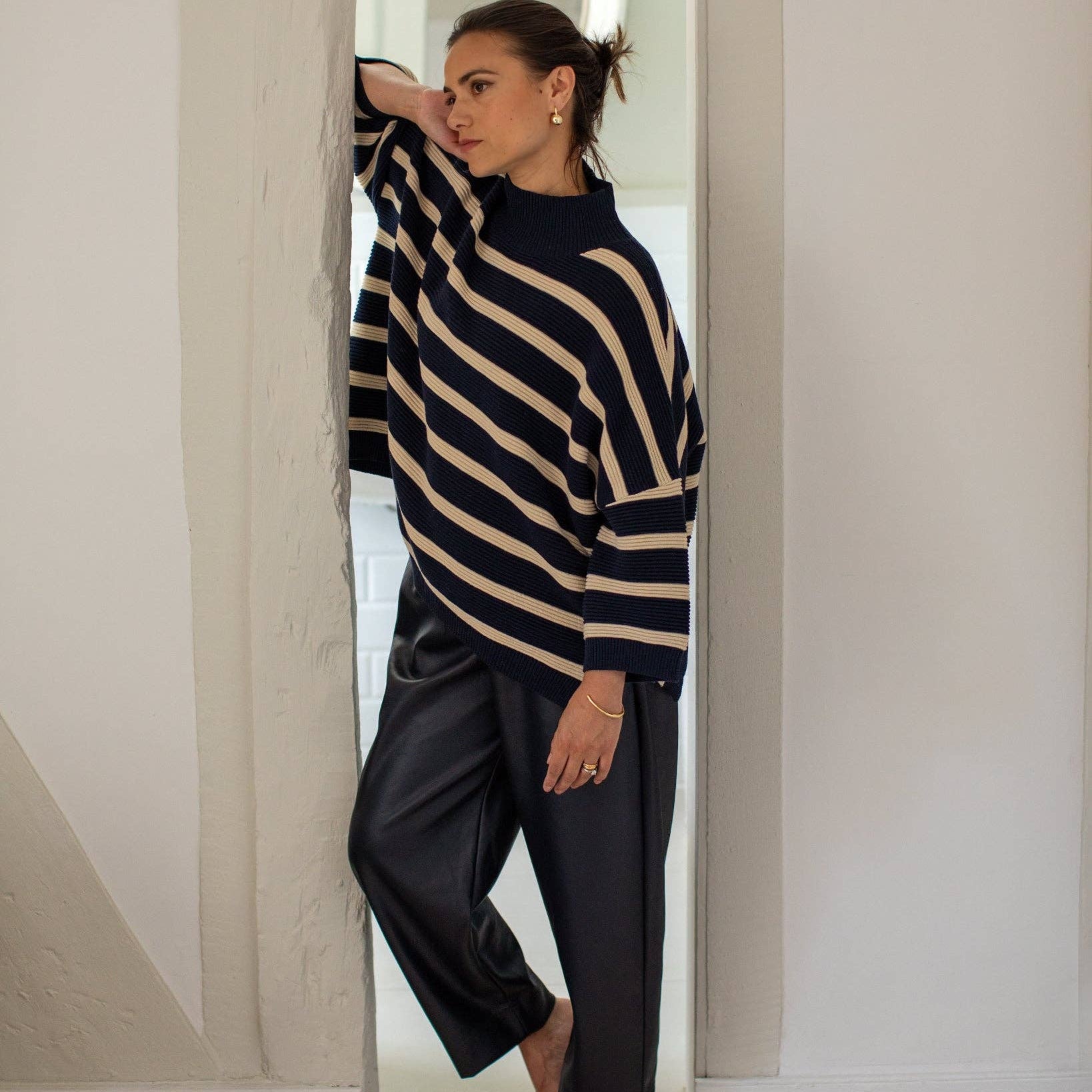 A woman stands barefoot in a doorway wearing a navy and beige striped knit sweater and dark wide-leg pants.