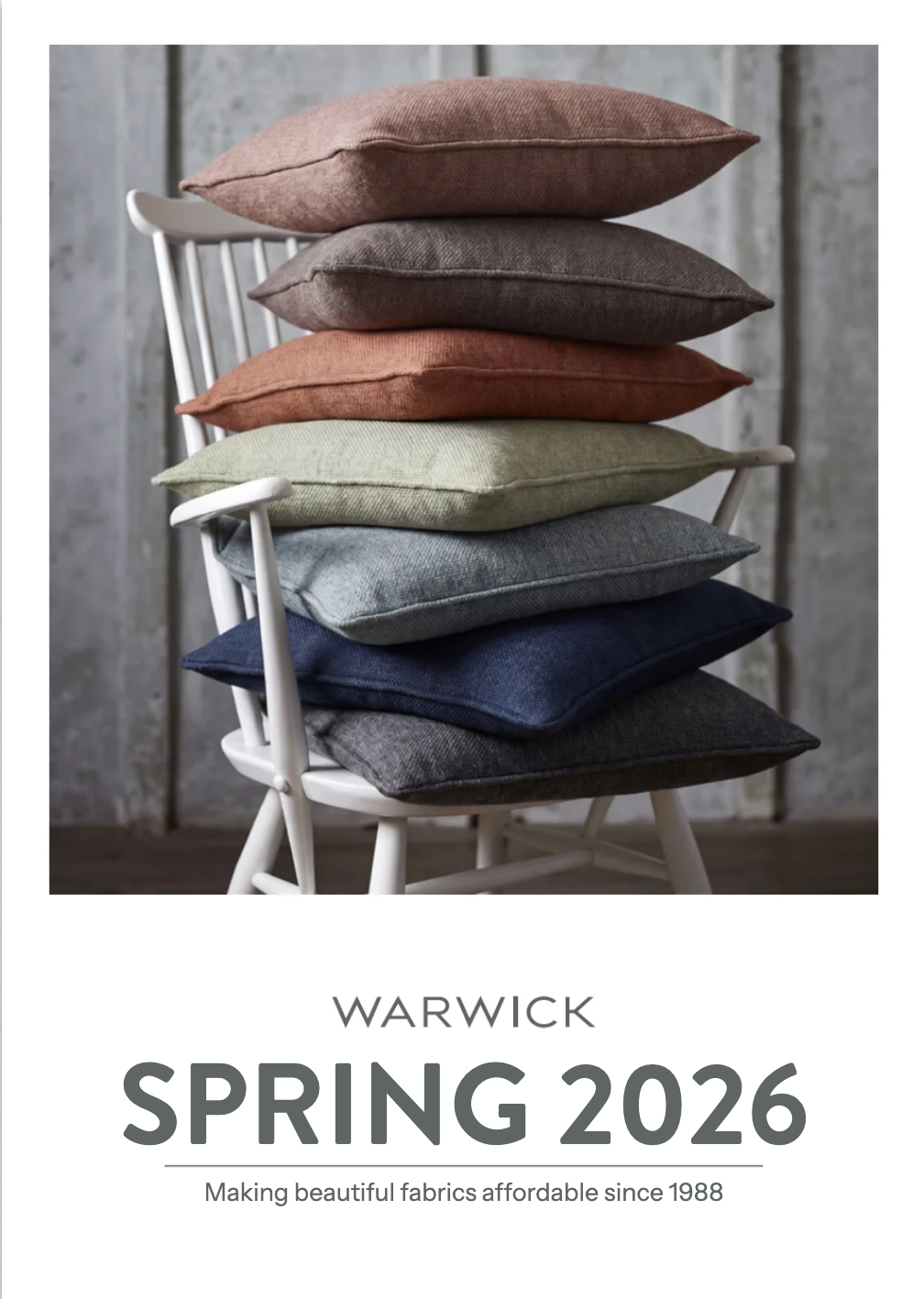 A catalog cover for Warwick Spring 2026, featuring a stack of seven textured throw pillows in muted earthy and cool tones resting on a white wooden chair against a gray wood-paneled wall. The bottom of the cover features the text 'WARWICK SPRING 2026' and the tagline 'Making beautiful fabrics affordable since 1988'.