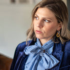A woman with shoulder-length brown hair wearing a navy velvet blazer over a blue and white striped blouse with a large bow, paired with gold hoop earrings, captured in a soft-focus indoor setting.