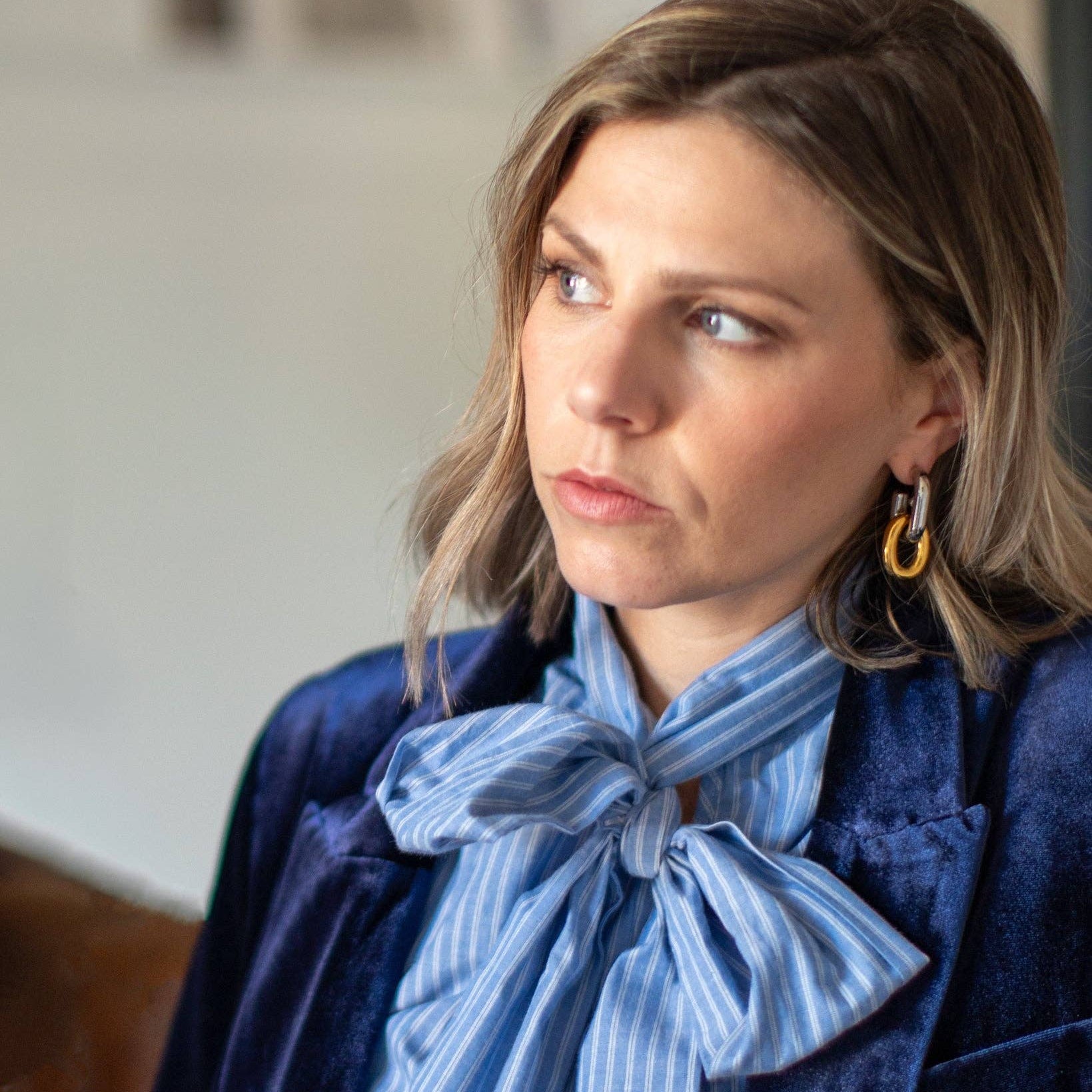 A woman with shoulder-length brown hair wearing a navy velvet blazer over a blue and white striped blouse with a large bow, paired with gold hoop earrings, captured in a soft-focus indoor setting.