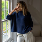 A woman with curly brown hair wearing a fuzzy navy blue mock-neck sweater and cream wide-leg trousers, standing by a window in a bright room.