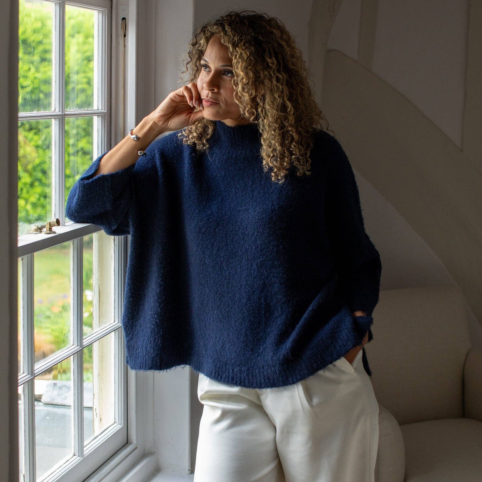 A woman with curly brown hair wearing a fuzzy navy blue mock-neck sweater and cream wide-leg trousers, standing by a window in a bright room.