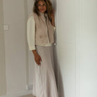 A woman stands in a minimalist white room wearing a beige faux-fur vest over a cream turtleneck and a light beige satin midi skirt, paired with black pointed flats.