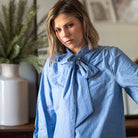 A woman wearing a light blue and white vertically striped blouse with a large bow tie-neck collar, standing in a softly lit indoor setting.