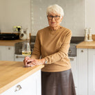 An older woman with short white hair and glasses stands in a bright kitchen, leaning on a wooden countertop with a glass of white wine. She wears a tan long-sleeve top and a dark brown skirt.