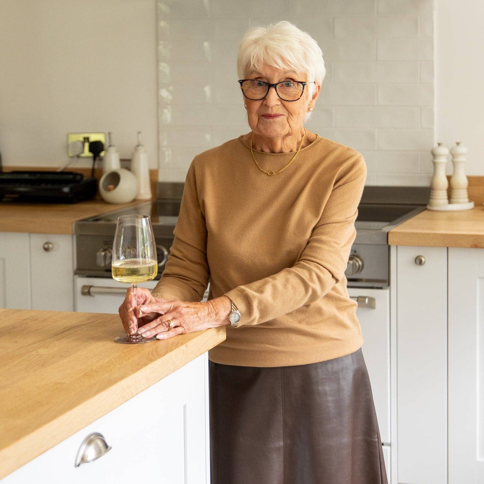 An older woman with short white hair and glasses stands in a bright kitchen, leaning on a wooden countertop with a glass of white wine. She wears a tan long-sleeve top and a dark brown skirt.