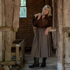 An older woman with blonde hair stands in a rustic wooden doorway of a brick building, wearing a dark brown patterned blouse, a long taupe skirt, and black boots.