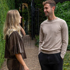 A man and a woman standing and talking on a gravel path in a garden setting. The man is wearing a light beige crewneck sweater and black trousers, while the woman wears a dark brown velvet puff-sleeve top and matching trousers.