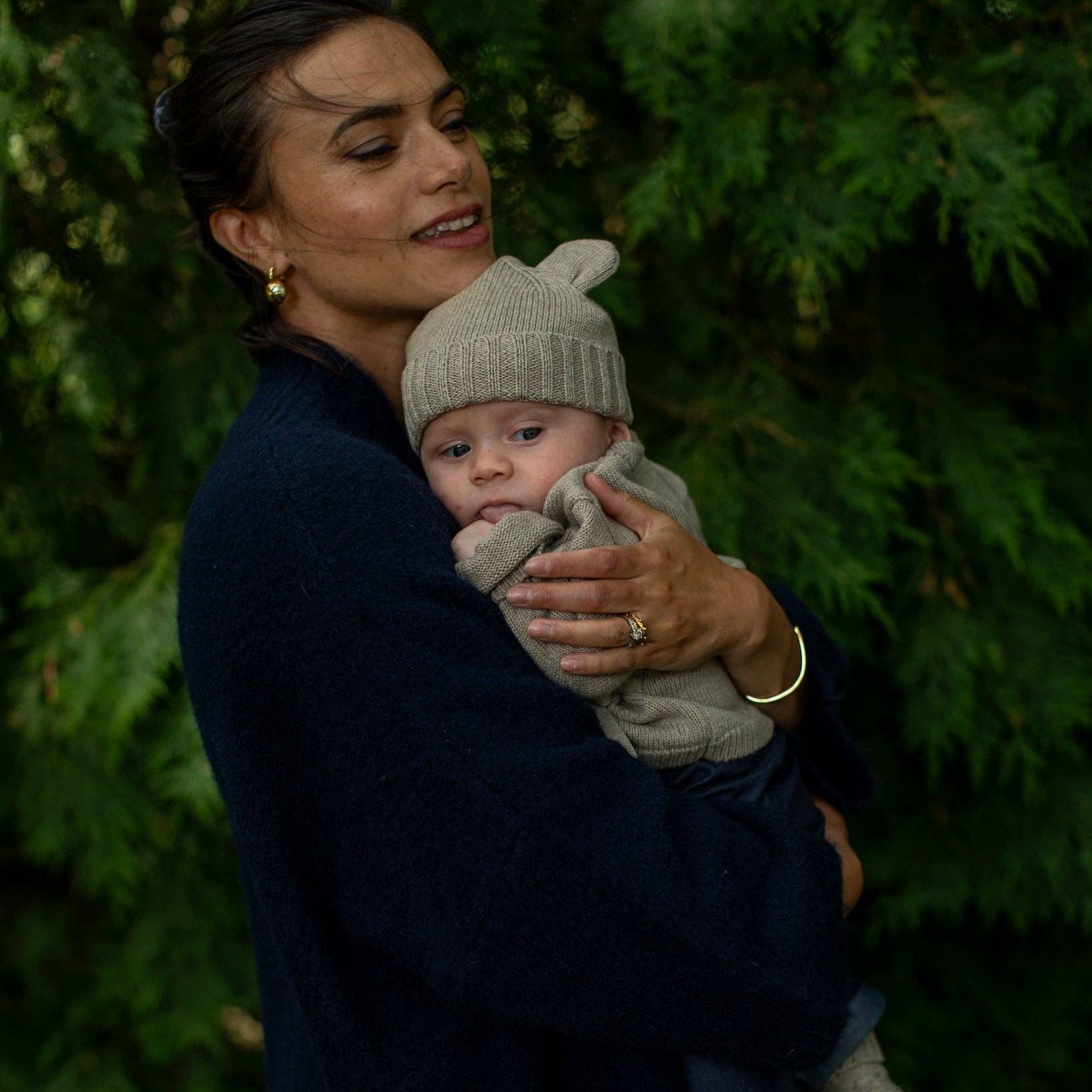 A woman in a dark navy knit cardigan holds a baby wearing a light beige knit beanie and matching knit outfit, set against a dark green foliage background.