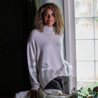 A woman with curly blonde hair wearing a cream knit mock-neck sweater and dark gray pants, leaning against a window in a dimly lit room with a pillow in the foreground.