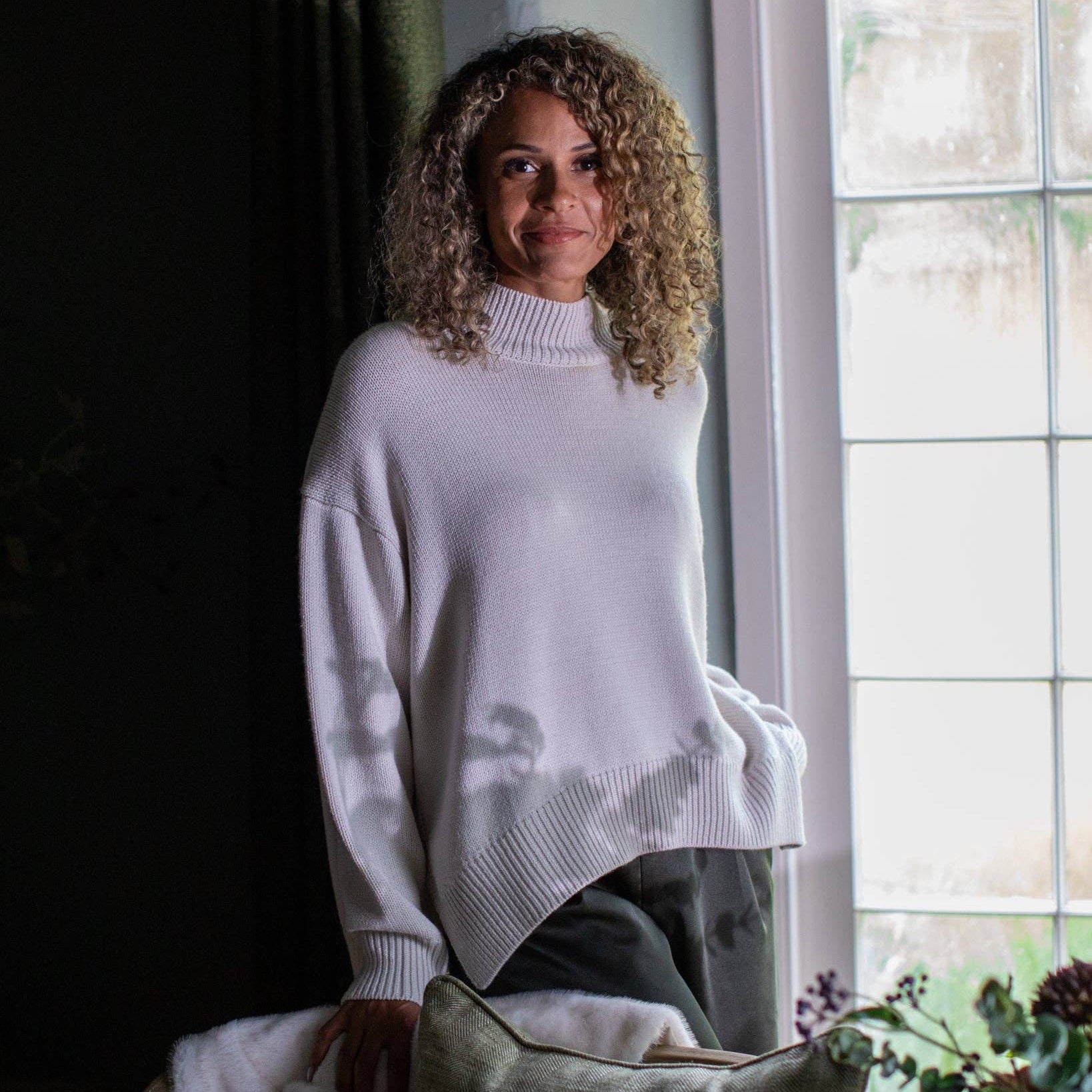 A woman with curly blonde hair wearing a cream knit mock-neck sweater and dark gray pants, leaning against a window in a dimly lit room with a pillow in the foreground.