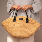 Person holding a straw bag with black handles against a neutral background