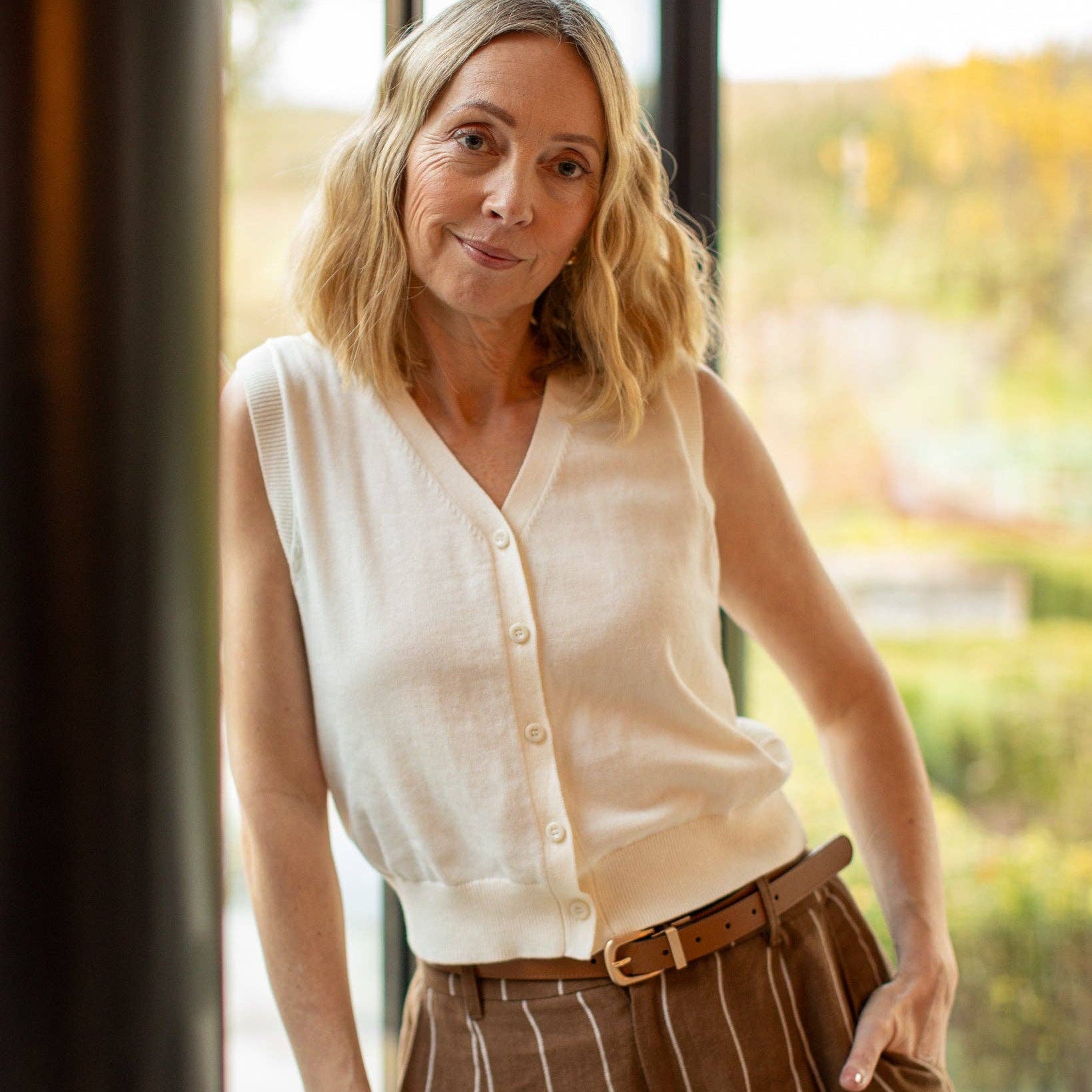 Woman wearing a white blouse and brown striped pants standing indoors with a blurred outdoor background