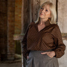 A woman with blonde hair stands in a rustic indoor setting, wearing a brown long-sleeve blouse with small white polka dots and a gray skirt.