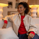 Woman sitting on a couch wearing a white jacket over a red shirt, with a neutral background.