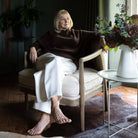 A woman sits barefoot in a cream upholstered armchair in a dark, stylish room. She is wearing a dark brown mock-neck knit sweater and white wide-leg trousers.