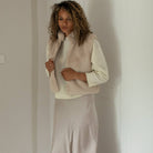 A woman with curly brown hair stands indoors wearing a cream long-sleeve top, a light beige faux-fur vest, and a matching beige satin skirt.