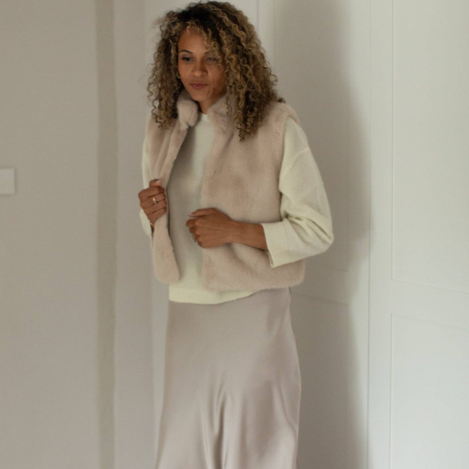 A woman with curly brown hair stands indoors wearing a cream long-sleeve top, a light beige faux-fur vest, and a matching beige satin skirt.