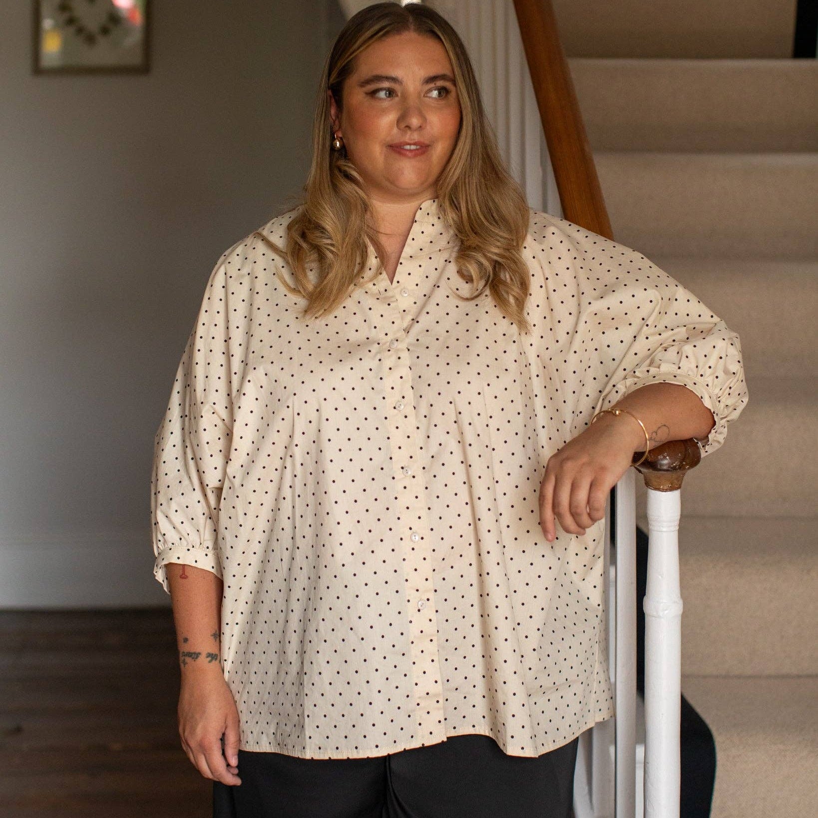A woman with blonde hair wearing a cream-colored, oversized 3/4 sleeve blouse with a small black polka dot pattern and black wide-leg pants, standing in a home interior next to a staircase.