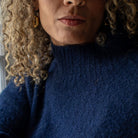 Close-up of a person wearing a textured navy blue knit turtleneck sweater, with curly blonde hair, gold hoop earrings, and a silver bangle visible.