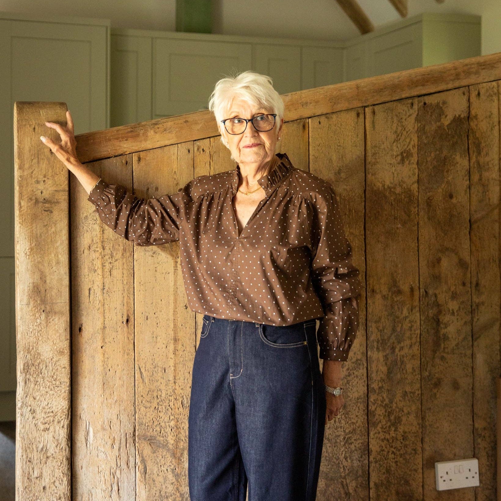 An older woman with short white hair and glasses stands indoors against a rustic wooden door, wearing a brown patterned blouse and blue jeans.