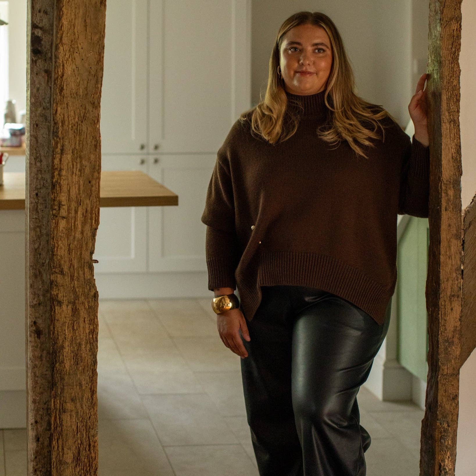 A woman wearing a brown turtleneck sweater and black wide-leg leather pants stands in a doorway of a modern kitchen.