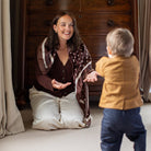 A woman kneels on a light beige carpet in a bedroom, smiling and gesturing toward a toddler standing with their back to the camera. The woman wears a brown cardigan over a dark brown top and cream-colored pants.