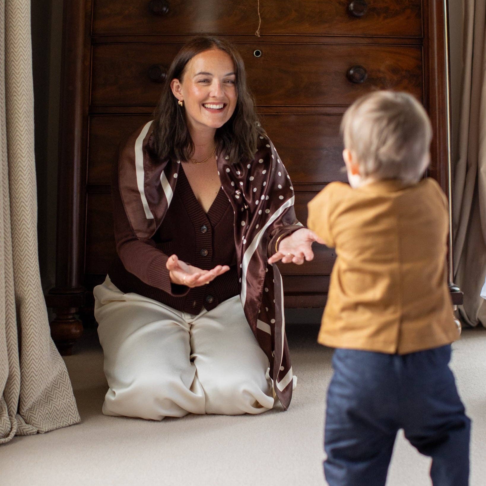 A woman kneels on a light beige carpet in a bedroom, smiling and gesturing toward a toddler standing with their back to the camera. The woman wears a brown cardigan over a dark brown top and cream-colored pants.