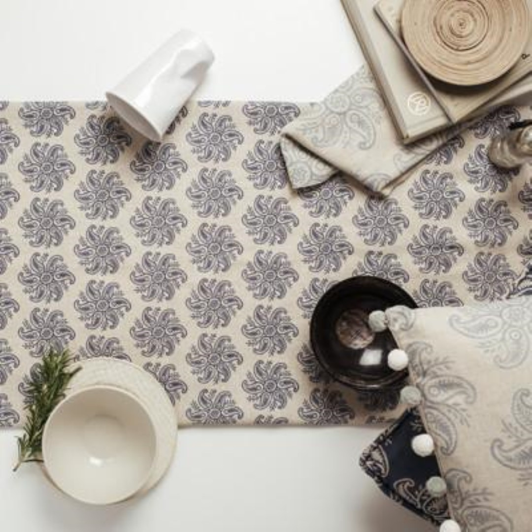 A table setting with a slate blue floral patterned fabric from Olive & Daisy, with a white bowl, wooden vase, and greenery decor.